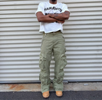 Person wearing a white t-shirt and green cargo pants standing in front of a corrugated metal wall.
